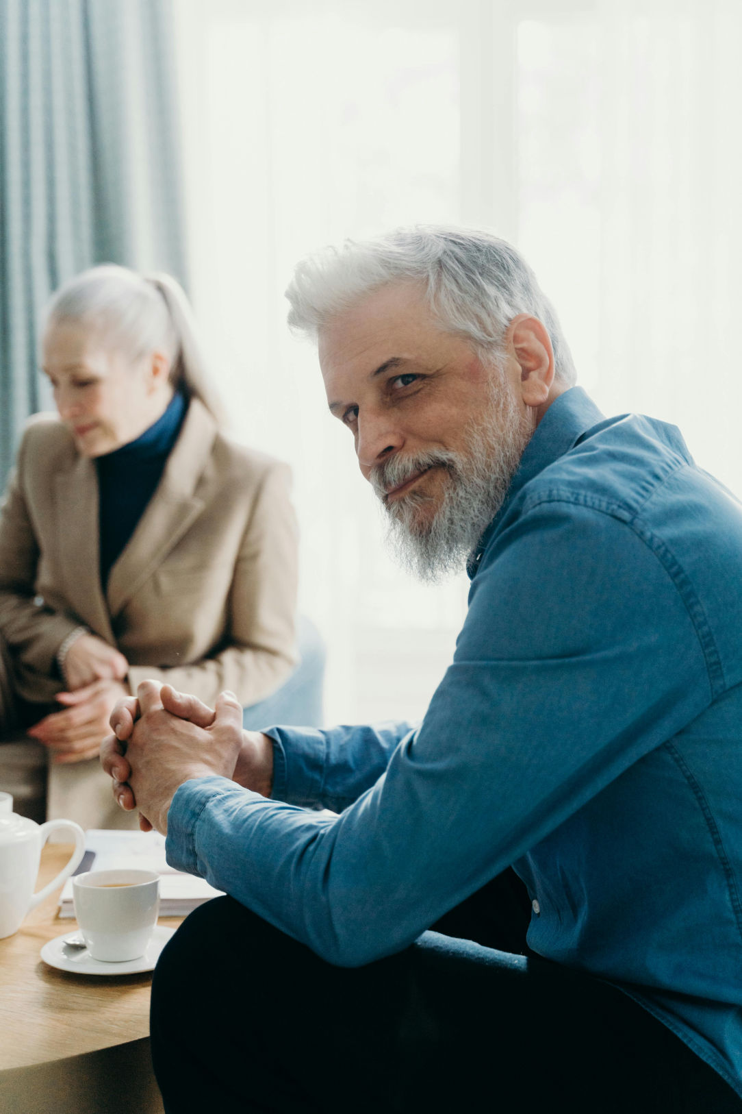 Hombre de mediana edad con barba y cabello canoso, vistiendo una camisa azul, sentado y sonriendo levemente mientras mira hacia la cámara. En el fondo, una mujer mayor con cabello recogido y abrigo beige parece concentrada en otra actividad. La escena transmite un ambiente relajado y elegante.