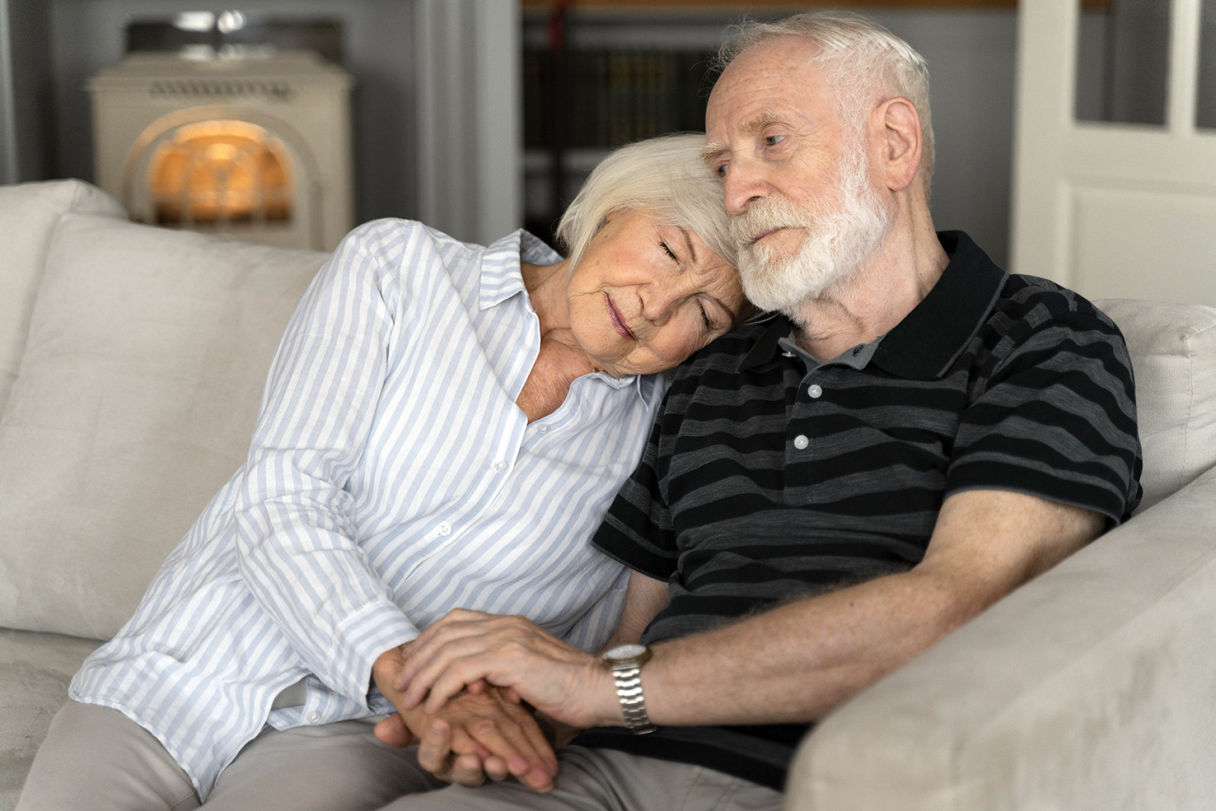 Pareja de personas mayores sentadas juntas en un sofá, abrazados y mirando hacia adelante. La mujer, con cabello corto y canoso, apoya su cabeza sobre el hombro del hombre, quien tiene barba y cabello gris. Ambos se sujetan de las manos, mostrando una expresión de ternura y complicidad.