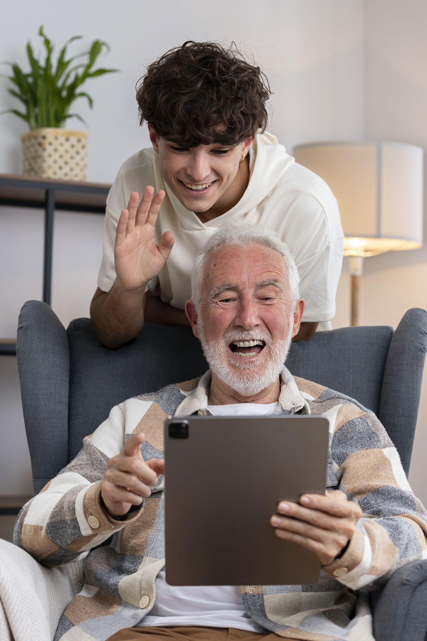 Hombre mayor con barba blanca y camisa de cuadros sonríe mientras usa una tableta digital, sentado en un sofá. Detrás de él, un joven con cabello rizado y sudadera blanca le acompaña, sonriendo y saludando con la mano. Ambos parecen estar disfrutando de una videollamada en un ambiente acogedor.